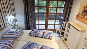 Ein Zimmer mit zwei Betten, einem Holztisch, einem Stuhl und einem Fenster mit Blick ins Grüne.
