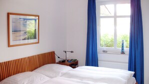 A bedroom with a bed, a nightstand, a lamp, a framed picture, and a window with blue curtains. - Kap Arkona by Interhome (Breege)