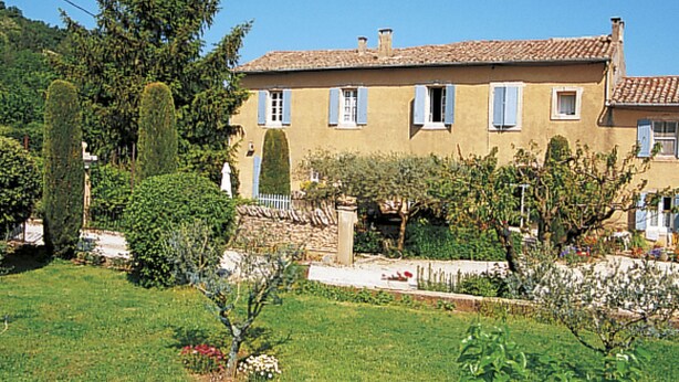 Une maison à deux étages, avec un toit en tuiles, entourée d’un jardin bien entretenu où poussent différentes espèces de plantes et où se trouve un petit étang.