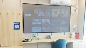 Ein Zimmer mit einem großen Flachbildfernseher an der Wand, einem Schreibtisch mit Stuhl, einer kleinen Mülleimer und einem Schild an der Wand.
