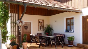 Una terraza de madera con techo de tejas, una mesa de comedor con sillas, plantas en macetas y una puerta de madera.