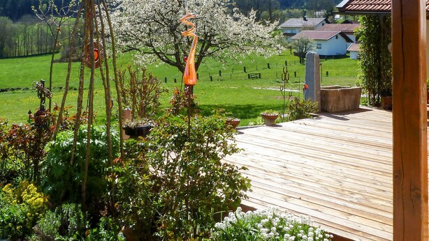Una terraza de madera con vistas a un jardín, una casa blanca y una colina boscosa al fondo.