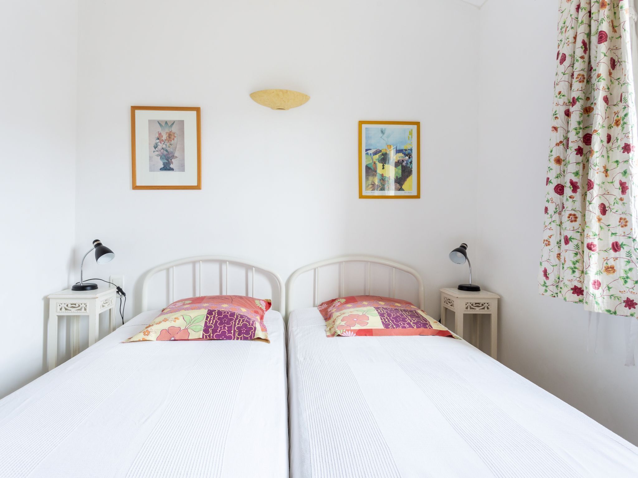 A bedroom with two beds, each with a white headboard and a colorful pillow. There are two bedside tables with lamps, and two framed pictures on the wall.
