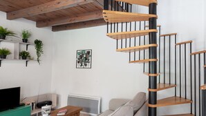 Moderni olohuone, jossa on kierreportaat, puupalkit, litteänäyttöinen televisio, sohva ja ruukkukasveja.