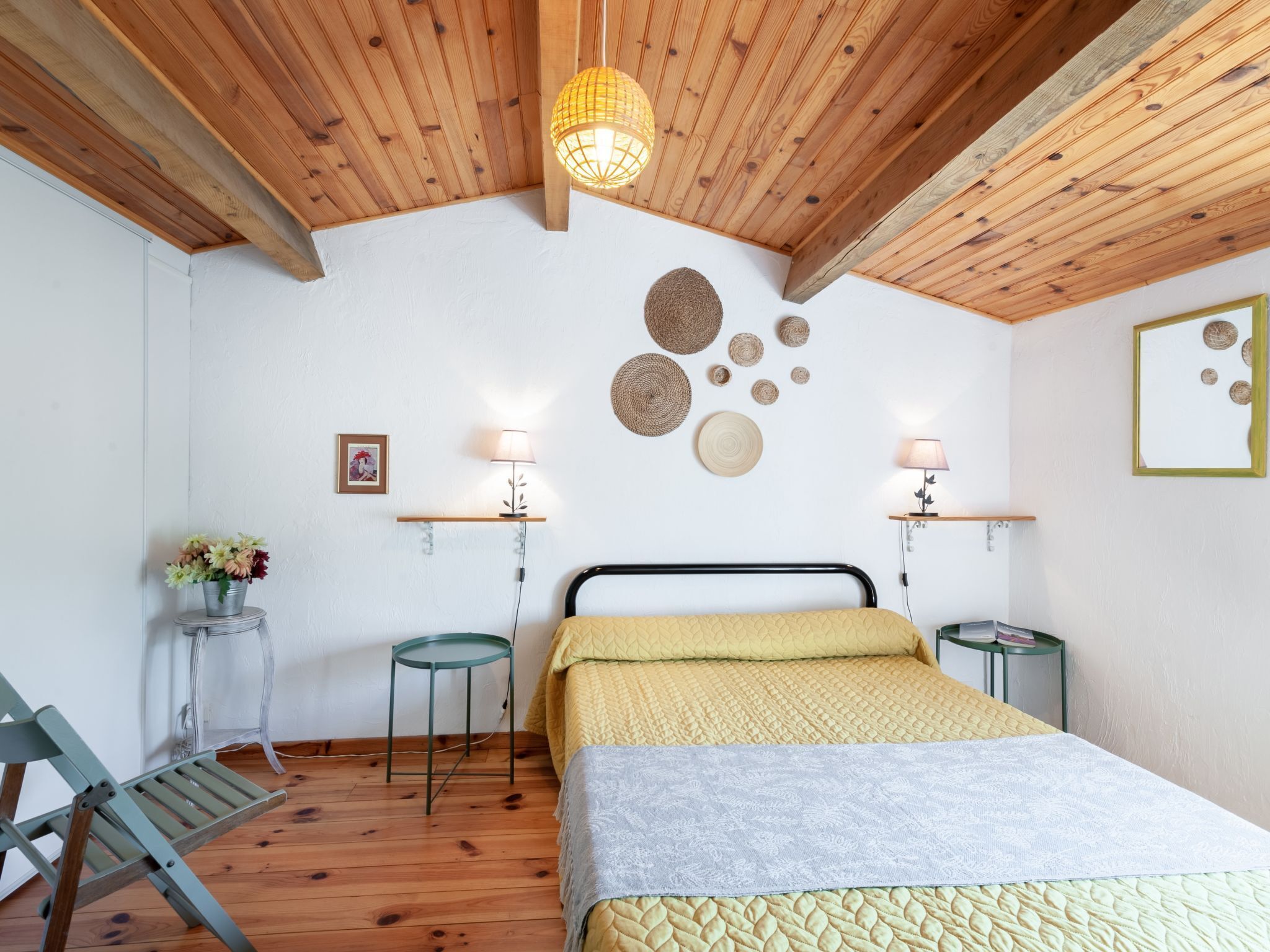 Een slaapkamer met een houten plafond, een bed met een gele sprei, twee nachtkastjes met lampen, een stoel en decoratieve wandkunst.