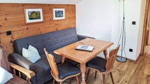 Uma sala de estar acolhedora com um sofá cinzento, mesa de jantar de madeira e cadeiras. Há quadros nas paredes e um abajur.