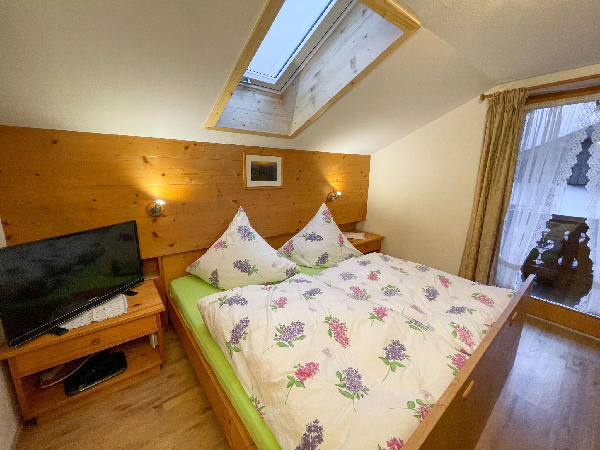 A bedroom with a wooden headboard, a bed with floral bedding, a television on a wooden stand, a bedside table, and a skylight.