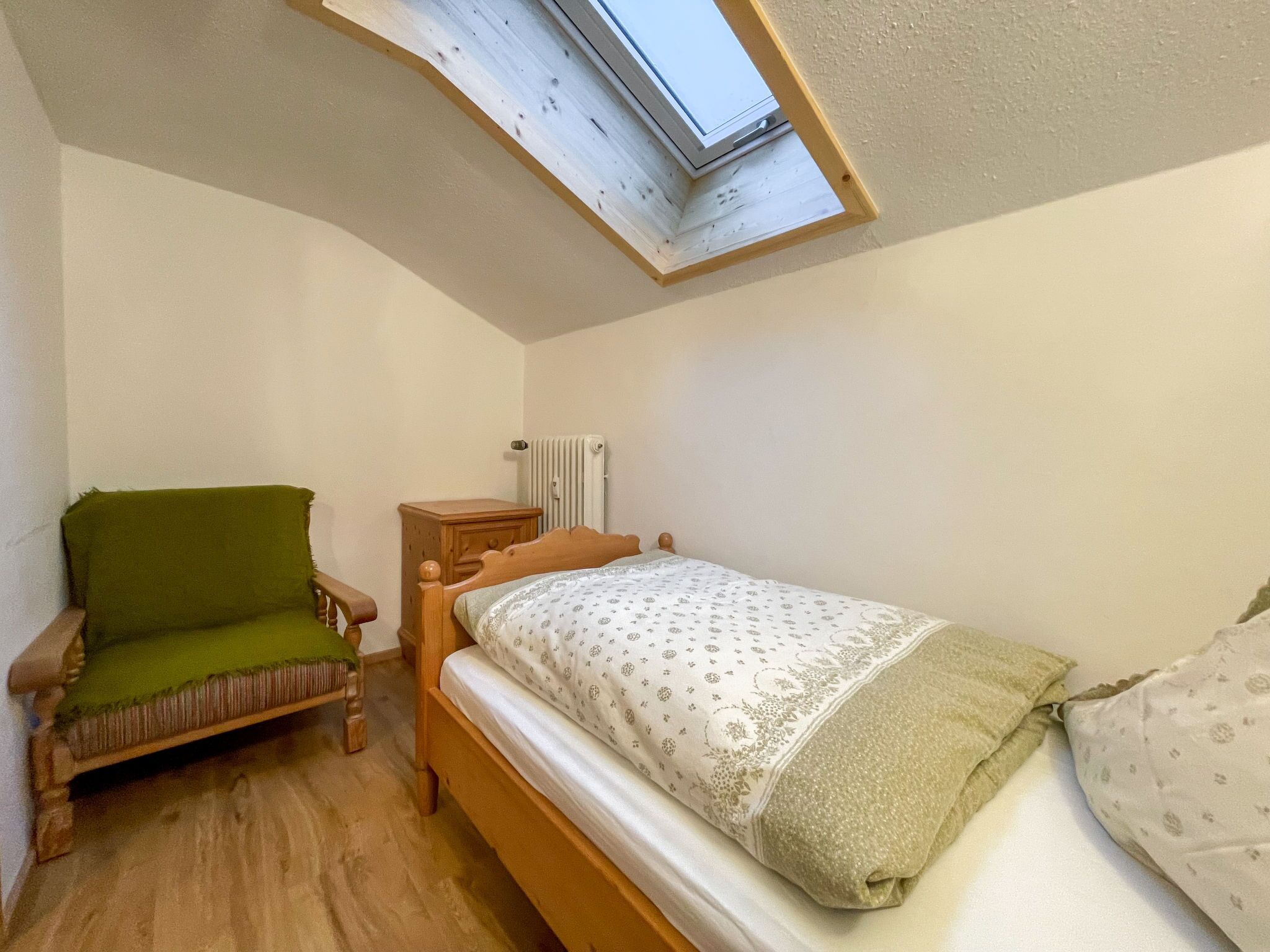 A small, cozy bedroom with a single bed, a wooden nightstand, a green chair, and a skylight above the bed.