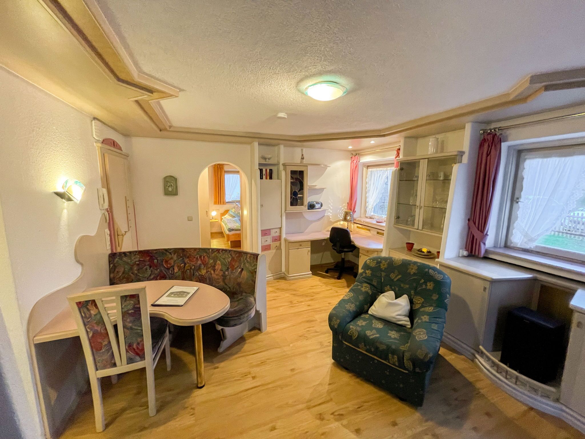 Ein gemütliches Zimmer mit Holzboden, einem kleinen runden Tisch, einem sofasessel mit Blumenmuster, einem einzelnen Sessel und einem Fenster mit Vorhängen.