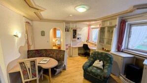 Un cuarto acogedor con piso de madera, una mesita redonda pequeña, un sofá estampado con motivos florales, un sillón individual y una ventana con cortinas.