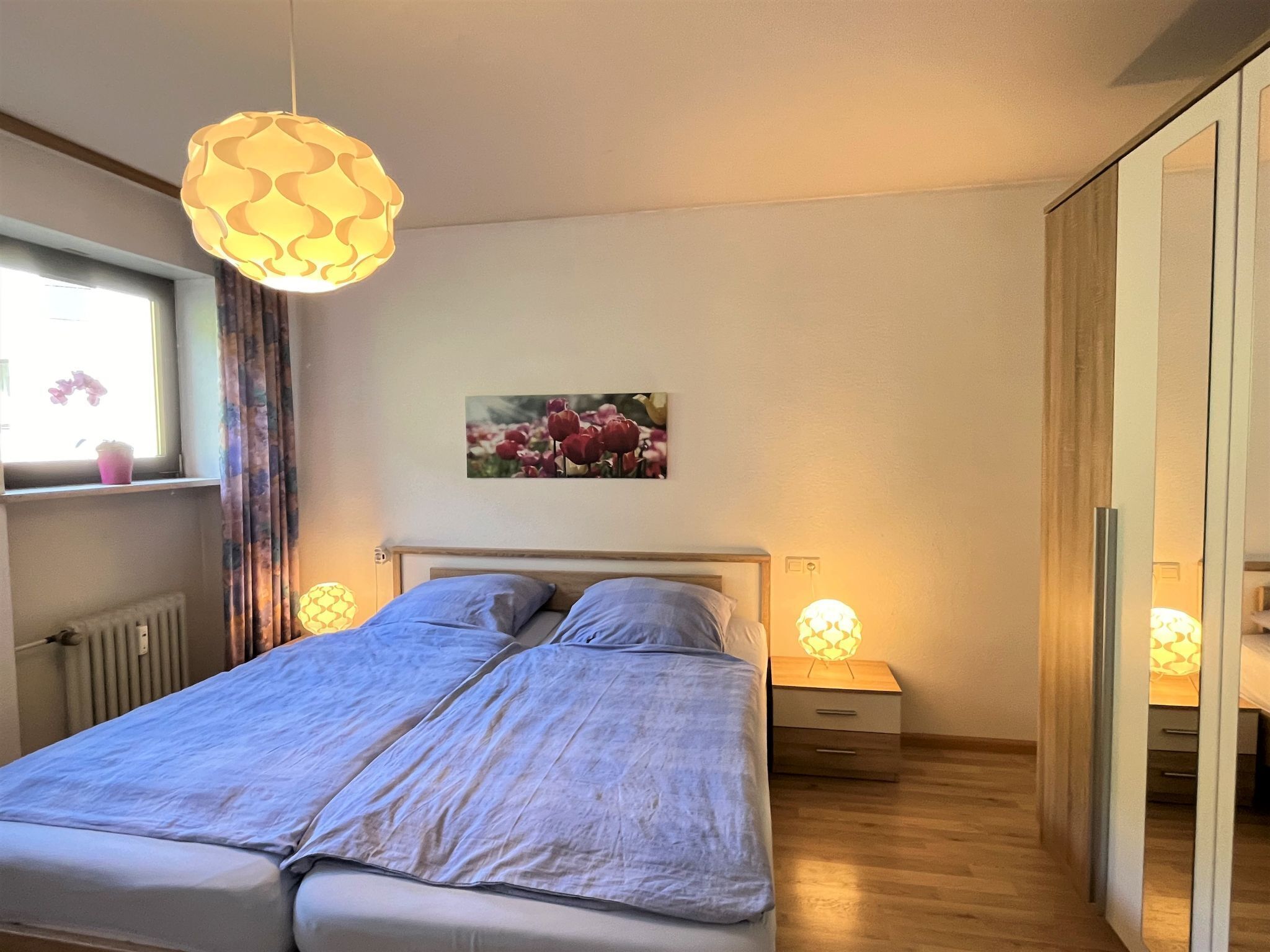 A bedroom with a bed, two bedside tables, a window with curtains, a radiator, a wall-mounted picture, and a wardrobe.