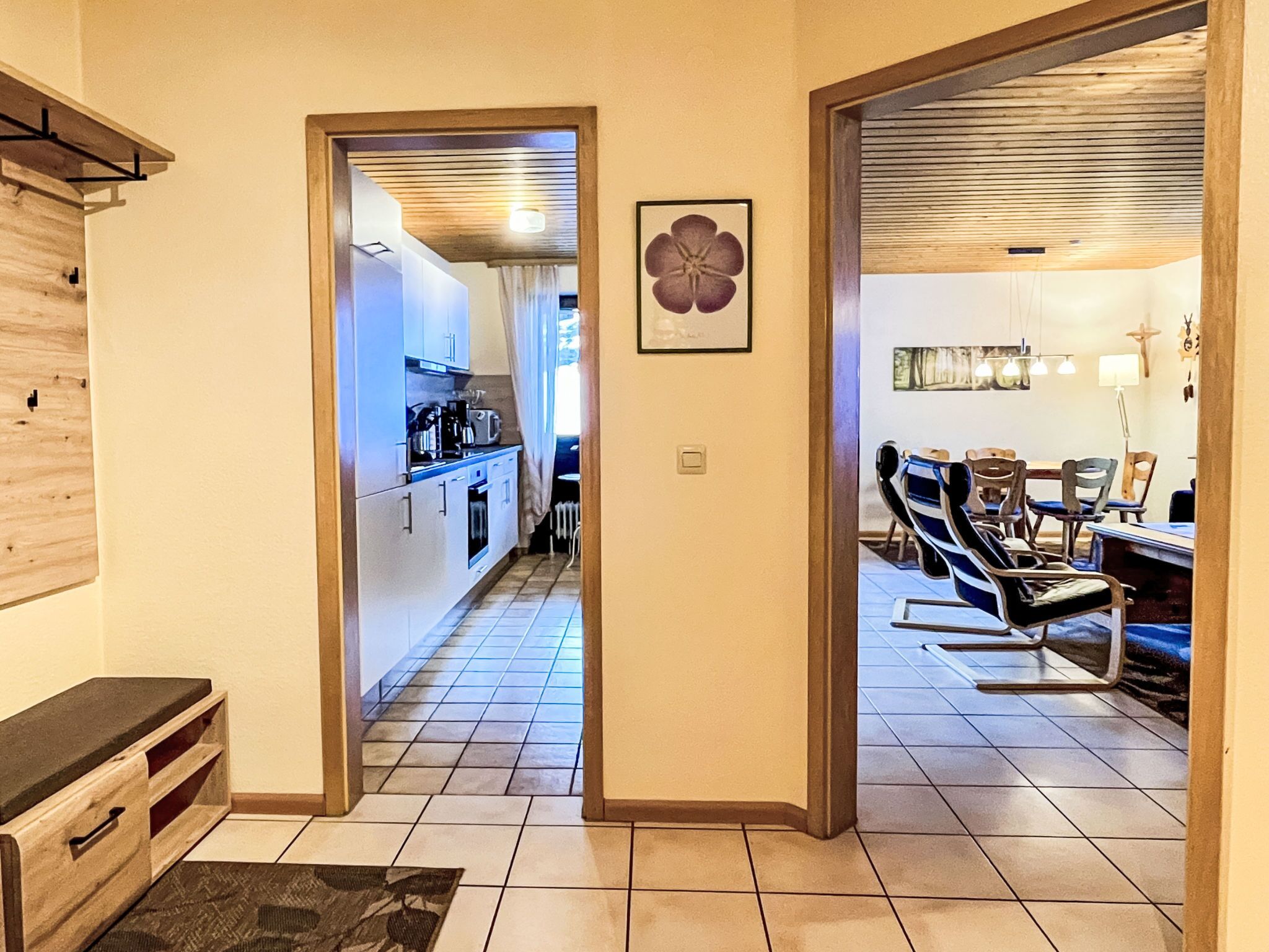 A hallway leading to a kitchen and dining area with wooden cabinets, a flower picture, and a dining table with chairs.