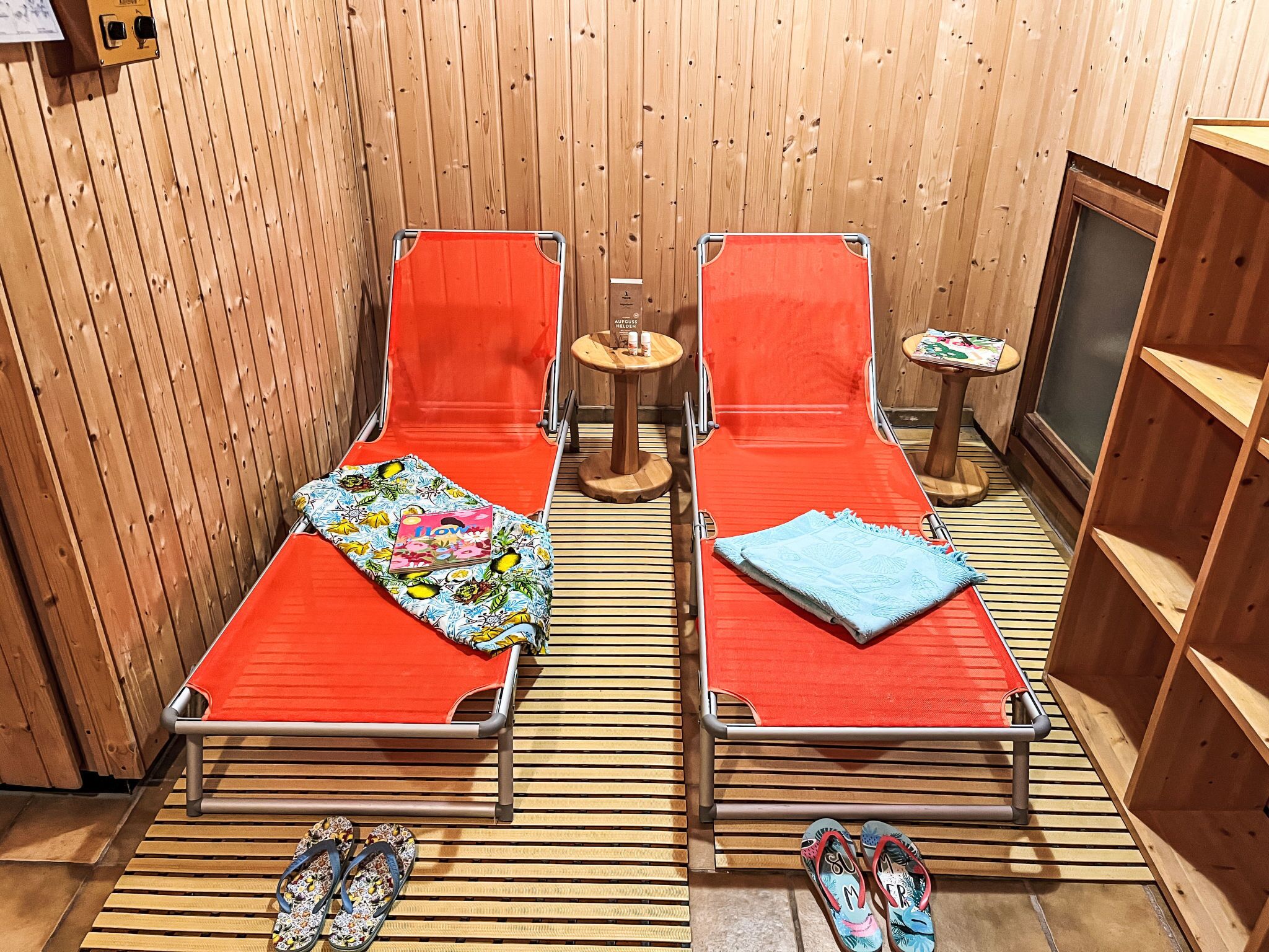 Two single beds with red mattresses and colorful blankets, a small table with a book, and a pair of flip-flops on the floor.