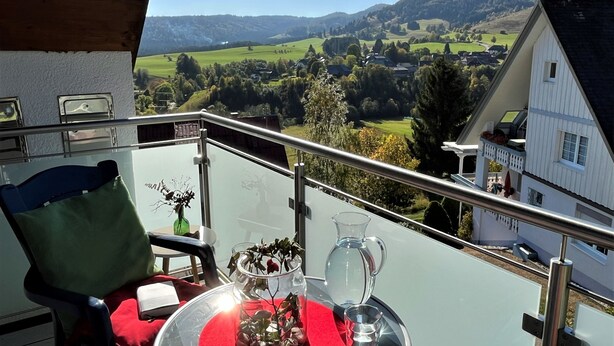 A balcony with a table set for two, a chair, and a view of a green valley and hills. - Schwarzwaldblick III by Interhome (Bernau)