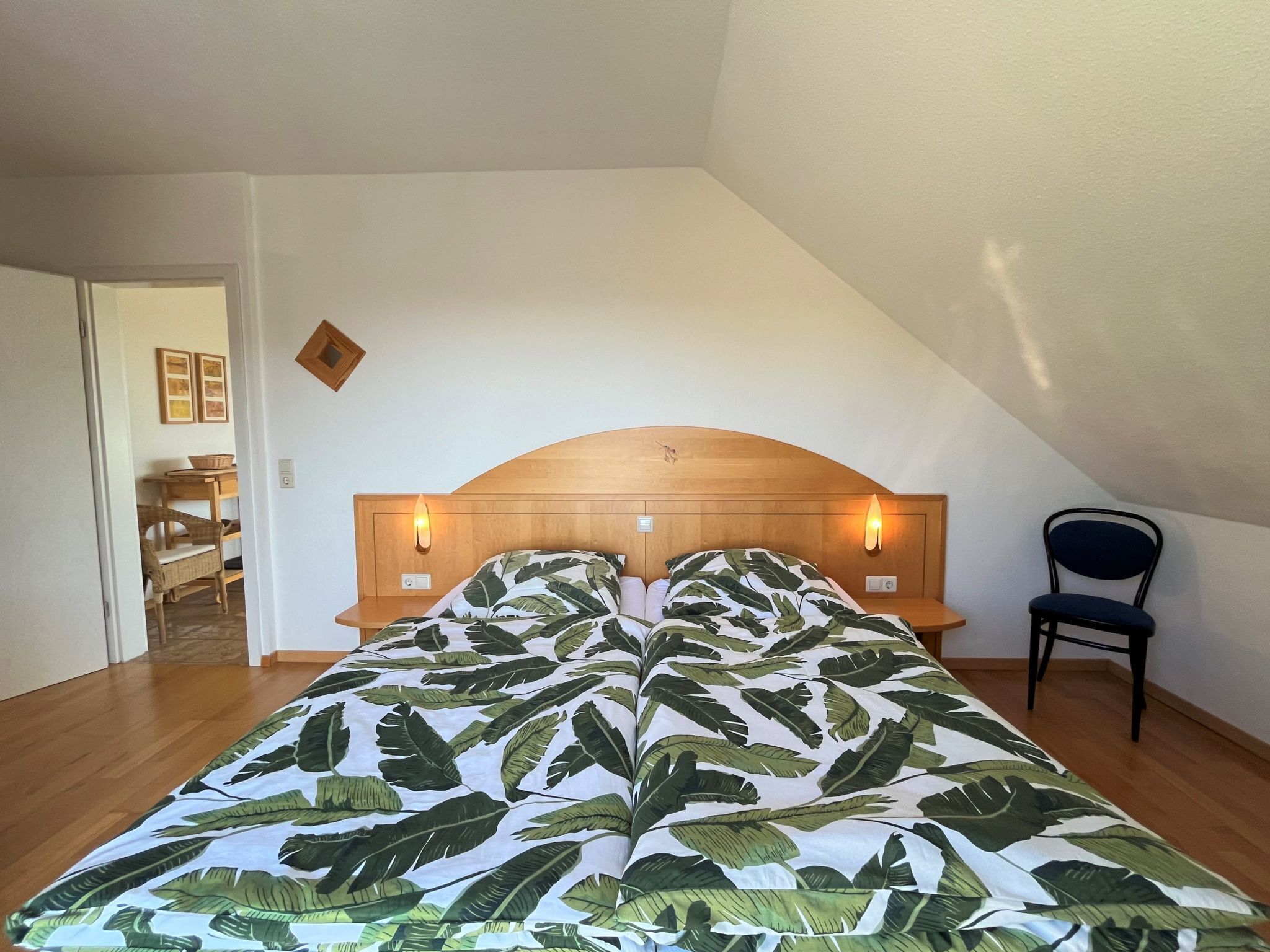 A bedroom with a sloped ceiling, a bed with a leaf-patterned bedspread, two wall-mounted reading lights, a chair, and a small table with a basket.