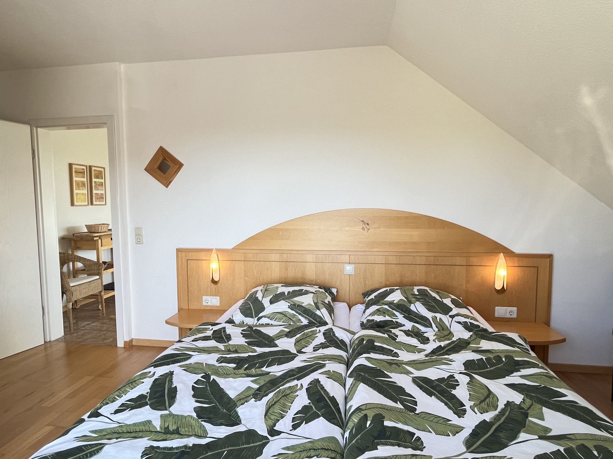 Une chambre à coucher avec une tête de lit en bois, deux lampes de chevet, un lit recouvert d’une couvre-lit à motifs de feuilles et une table de chevet en bois.