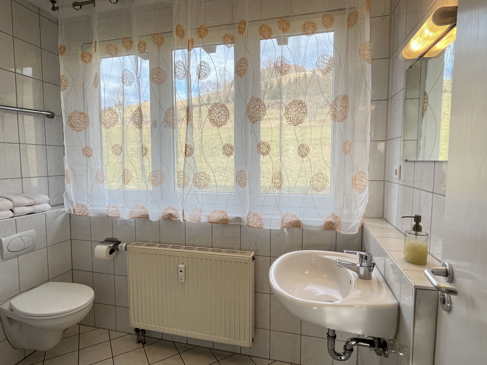 Une salle de bain comprenant des toilettes, un lavabo, un radiateur et une fenêtre agrémentée de rideaux à motifs floraux.