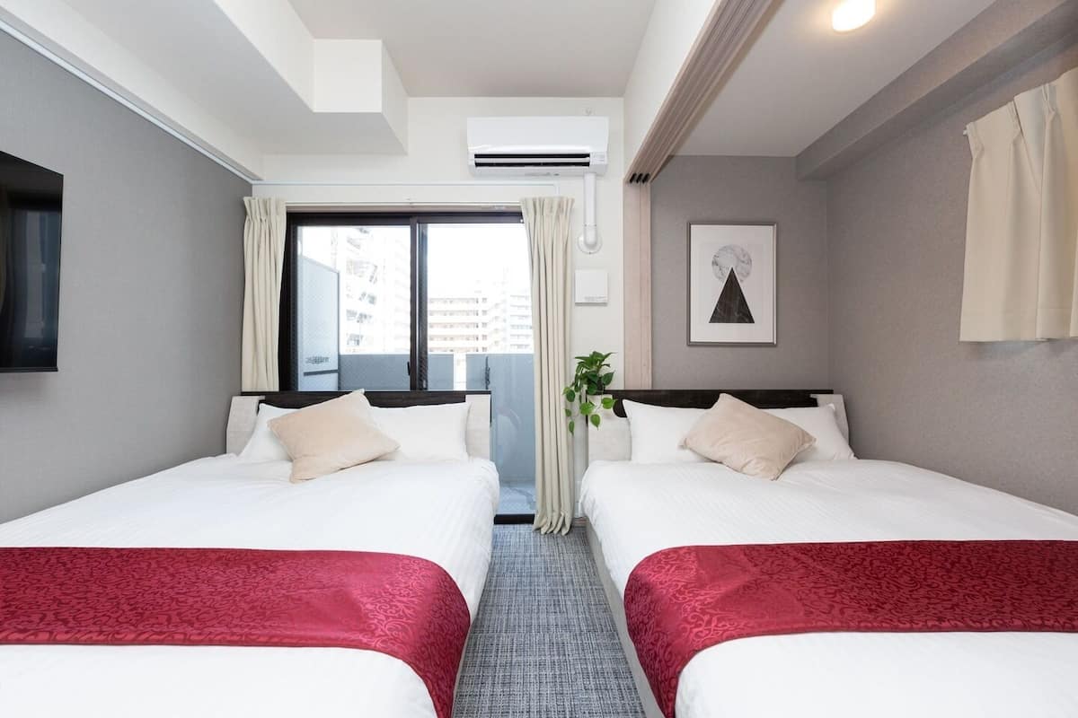 Habitación de hotel con dos camas, una ventana con cortinas, un aire acondicionado, un cuadro en la pared y una planta.