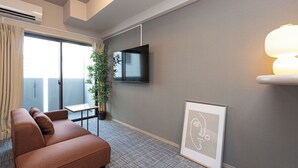 Ein modernes Hotelzimmer mit Sofa, Flachbildfernseher an der Wand, Zimmerpflanze und einem gerahmten Kunstwerk.