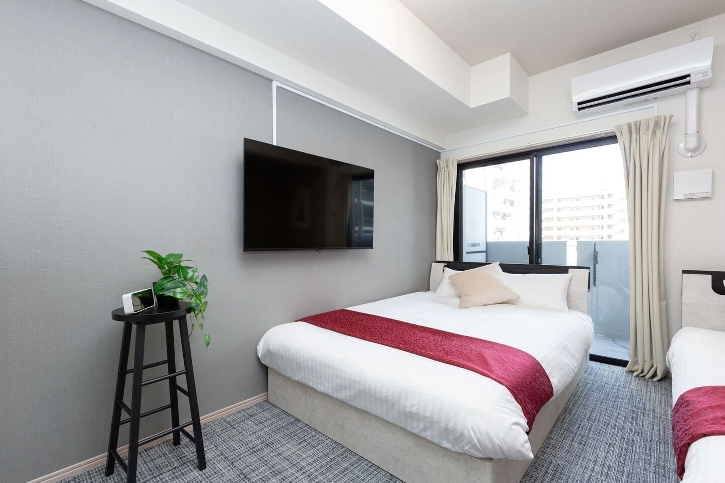 A hotel room with a bed, a TV mounted on the wall, a small table with a plant, and a window with curtains.
