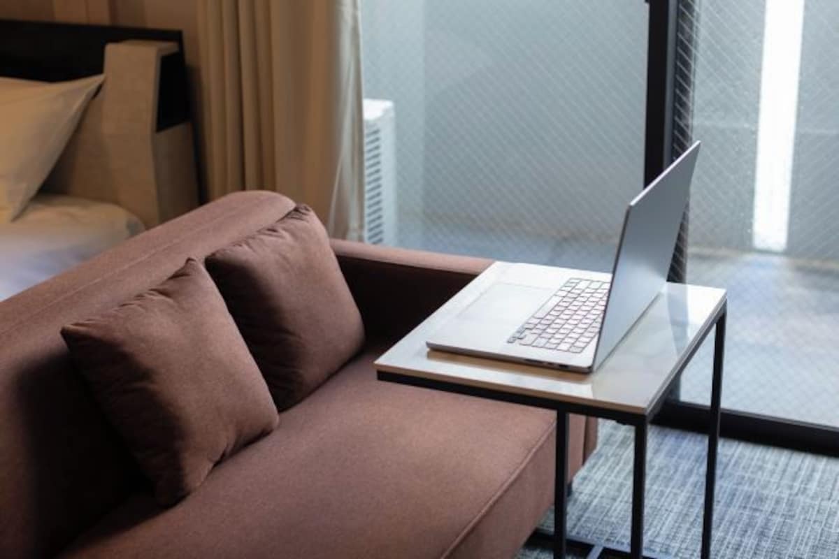 Quarto de hotel moderno com um sofá marrom, um laptop sobre uma mesinha e vista da cidade através de amplas janelas.