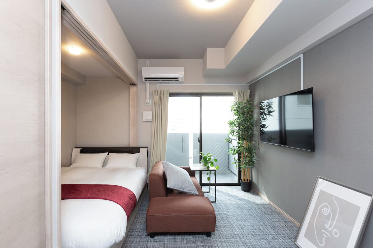 Habitación de hotel con cama, sofá, televisor, planta y balcón con vistas.