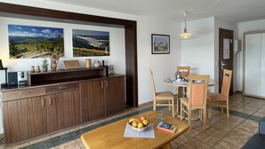 A dining area with a wooden table and chairs, a small kitchen area with cabinets and a countertop, and two framed landscape paintings on the wall.