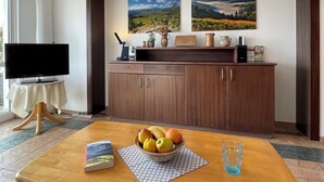 A living room with a TV, wooden cabinet, two paintings, a bowl of fruit, a book, and a glass of water on a table.