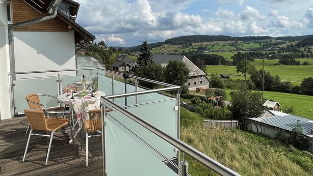 A balcony with a table and chairs, overlooking a scenic landscape with houses and fields.
