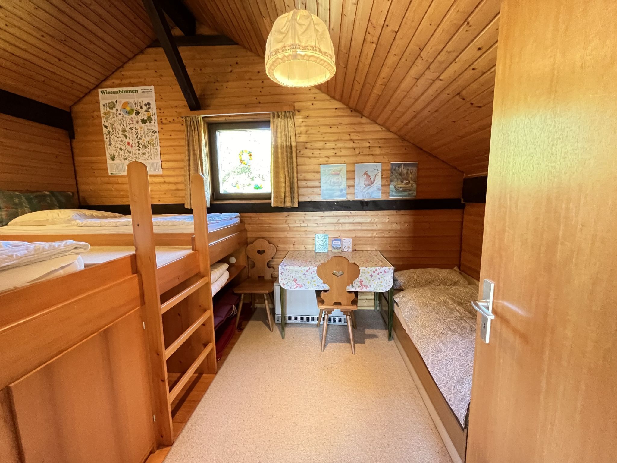 A cozy attic bedroom with wooden walls and a sloped ceiling. It features two bunk beds, a small desk with a chair, and a window with curtains.