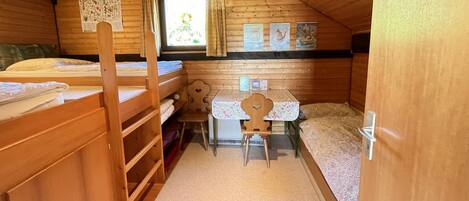 A cozy attic bedroom with wooden walls and a sloped ceiling. It features two bunk beds, a small desk with a chair, and a window with curtains.