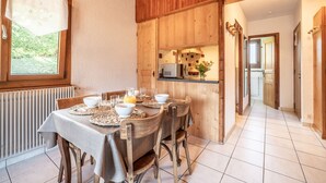 Zona pranzo con tavolo in legno apparecchiato per quattro, una finestra con vista sul verde e una cucina visibile attraverso una porta aperta.