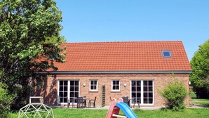 Un bâtiment en briques surmonté d’un toit de tuiles rouges, une aire de jeux avec un toboggan et une petite table avec des chaises dans la cour.