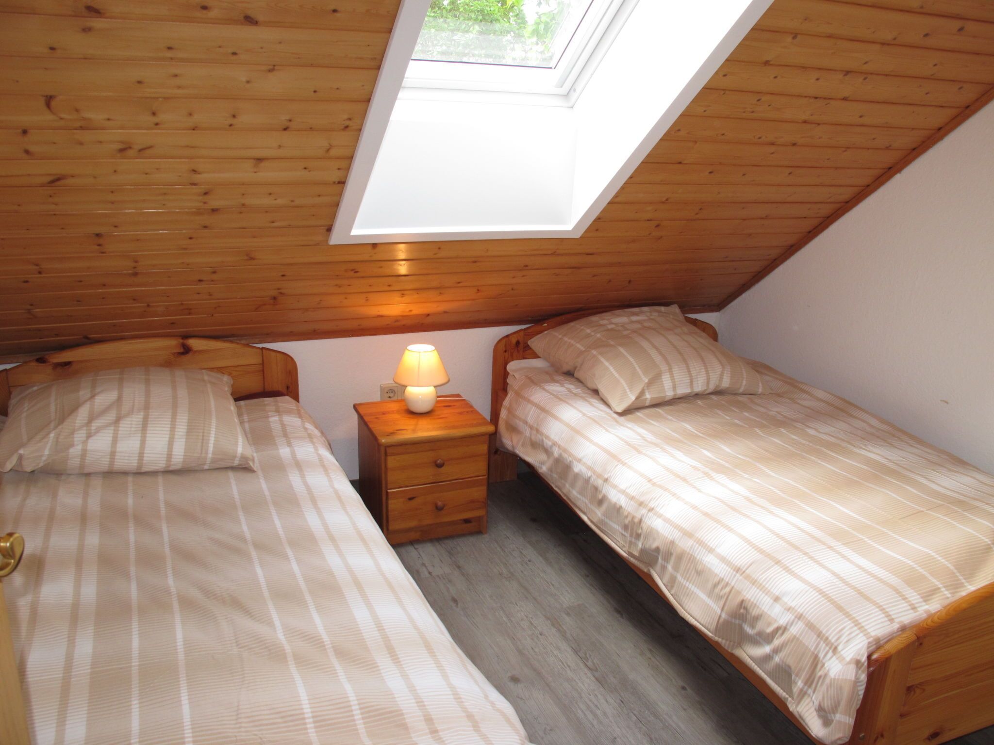 A small, cozy room with two wooden beds, a nightstand with a lamp, and a skylight above.