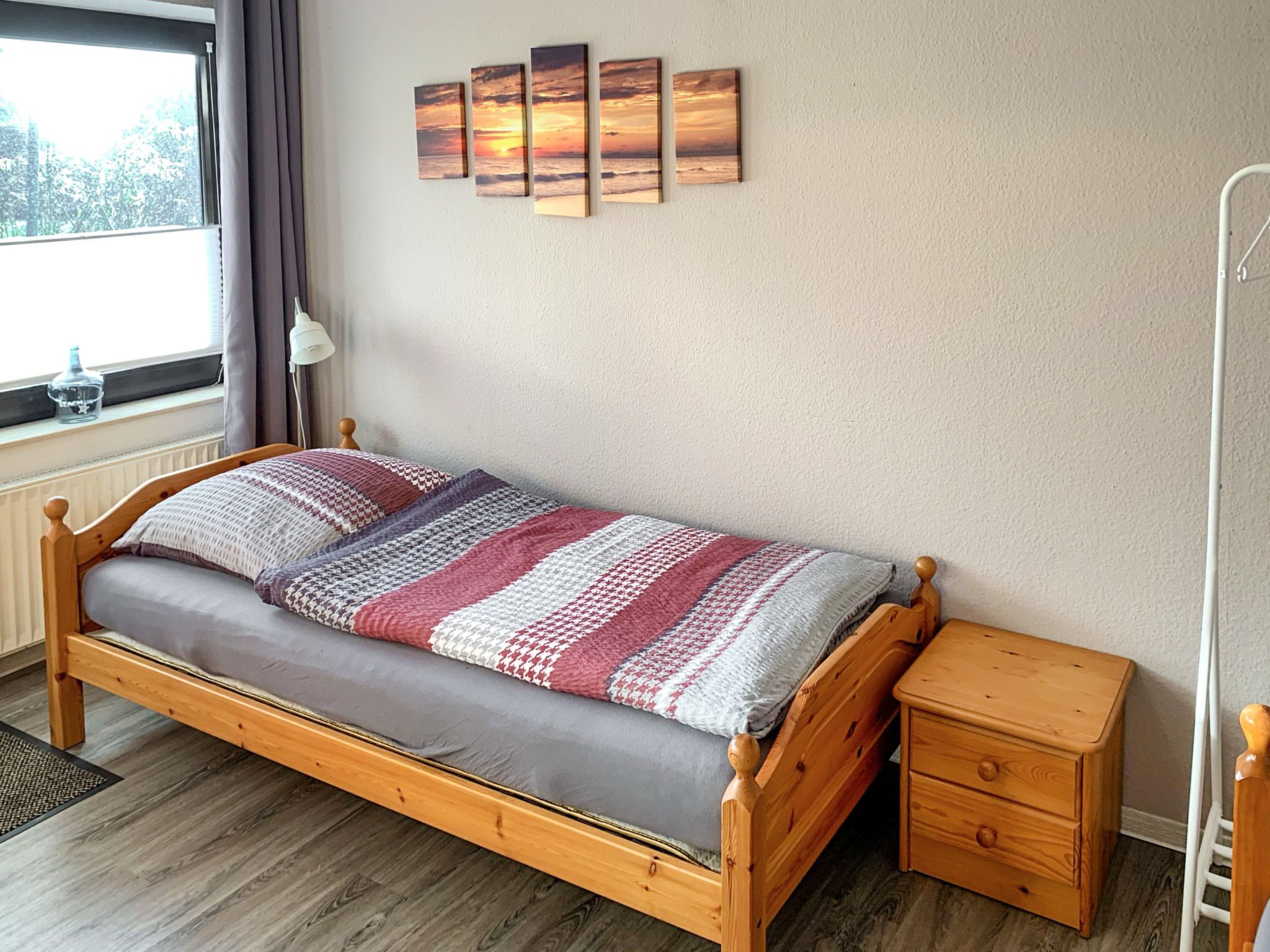 A single bed with a wooden frame and a gray mattress, a bedside table with drawers, a wall-mounted shelf with four framed pictures, and a window with curtains.