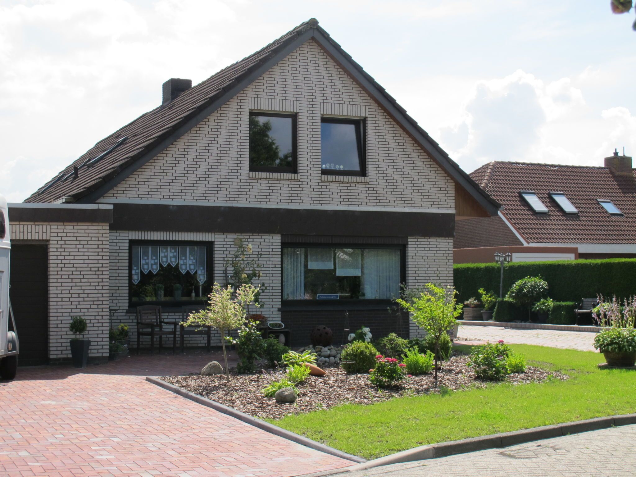 A two-story house with a brick facade, a pitched roof, and a front garden with various plants and a paved driveway.