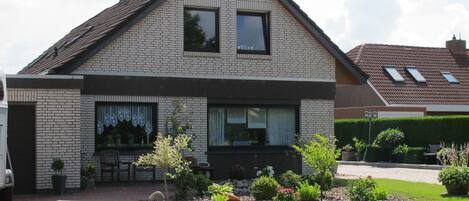 A two-story house with a brick facade, a pitched roof, and a front garden with various plants and a paved driveway.