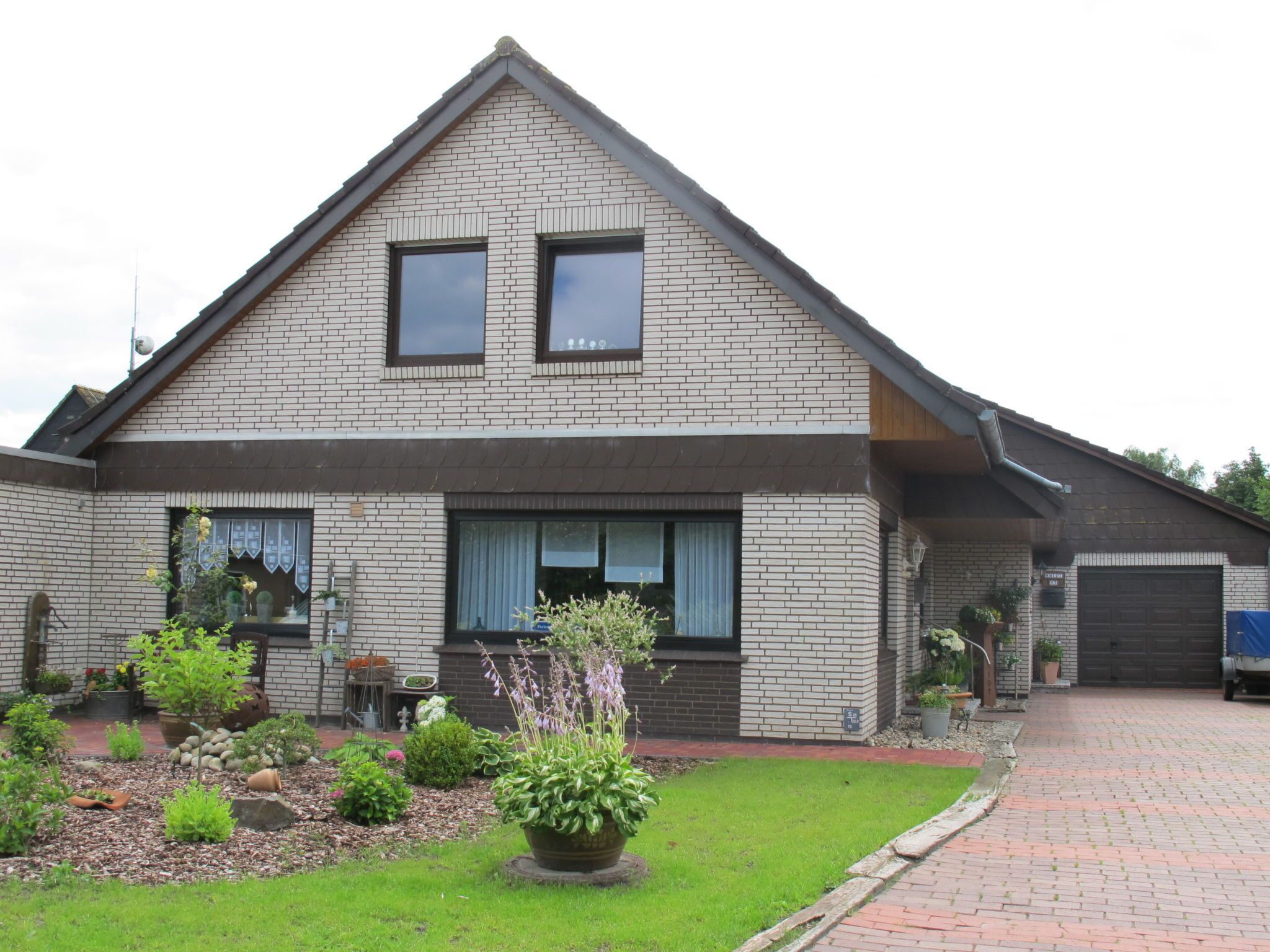 A two-story house with a brick exterior, a sloped roof, and a front garden with various plants and a lawn.