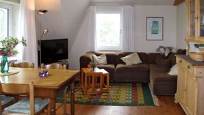 A cozy living room with a wooden dining table, a sofa, a television, and a window with curtains. - Landhaus Mahnhamm by Interhome (Horumersiel)