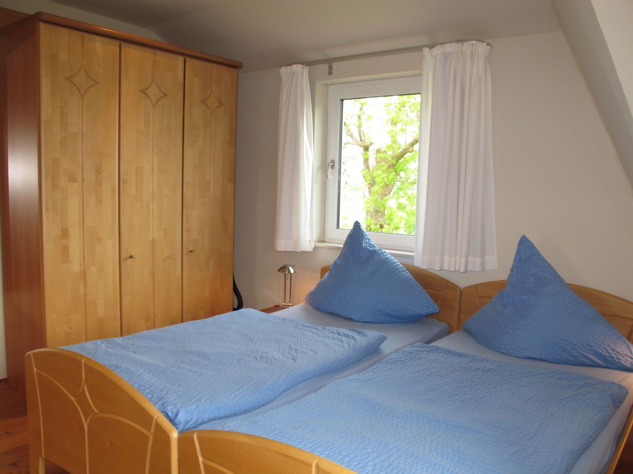 Quarto com duas camas de solteiro, um guarda-roupa de madeira, uma janela com cortinas e uma mesa de cabeceira com abajur.