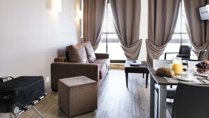 Ein modernes Hotelzimmer mit einer Couch, Hocker, Esstisch und einem Koffer.
