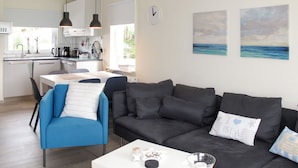 A modern living room with a black sofa, a blue armchair, and a white coffee table. There is a kitchen in the background with white cabinets and a window. - Tossens by Interhome (Tossens)