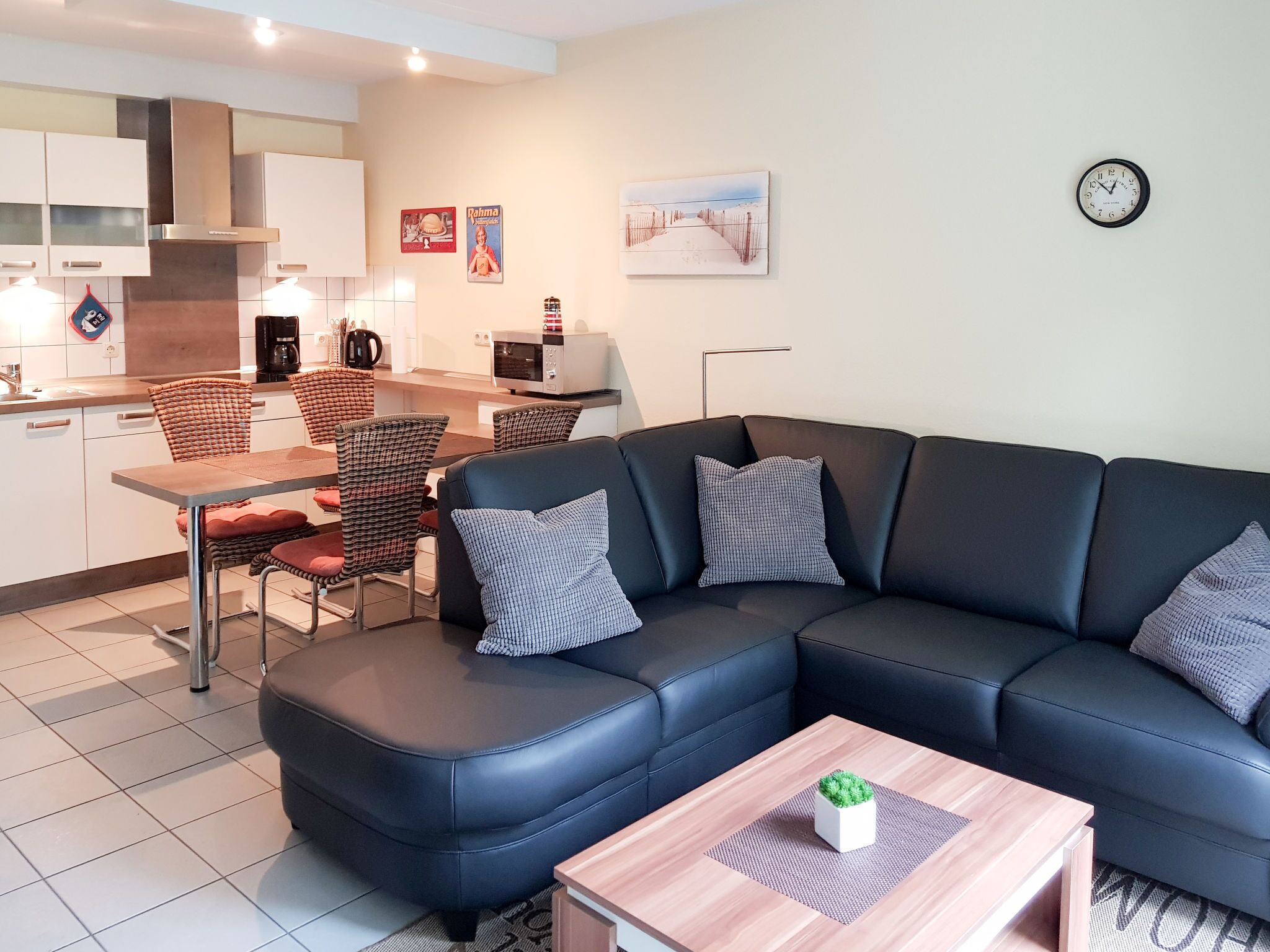 A modern living room with a sectional sofa, a dining area with wicker chairs, a kitchen with white cabinets, and a clock on the wall.