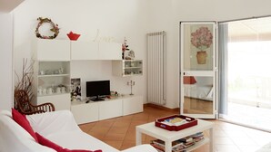 A modern living room with a white sofa, a coffee table, and a TV. There is a bookshelf with decorative items and a glass door leading to an outdoor area. - Villa Veranda by Interhome (Sanremo)