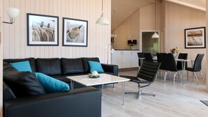 A modern living room with a black leather sofa, a small table, and a dining area with black chairs. There are framed pictures on the wall, a kitchen in the background, and wooden paneling on the walls. - Holiday Vital Resort (GBE111) by Interhome (Großenbrode)