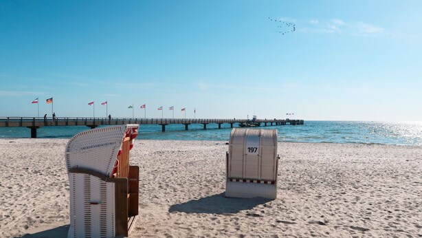 A beach with two wicker chairs and a numbered bench, a pier with flags, and a clear sky. - Holiday Vital Resort (GBE111) by Interhome (Großenbrode)