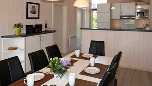 Une cuisine moderne avec des meubles blancs, une table à manger dressée avec des assiettes, des tasses et un centre de table. Des luminaires suspendus sont placés au-dessus de la table, et une fenêtre apporte une lumière naturelle.
