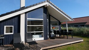 Een modern huis met een groot raam, een terras met stoelen en een schoorsteen.