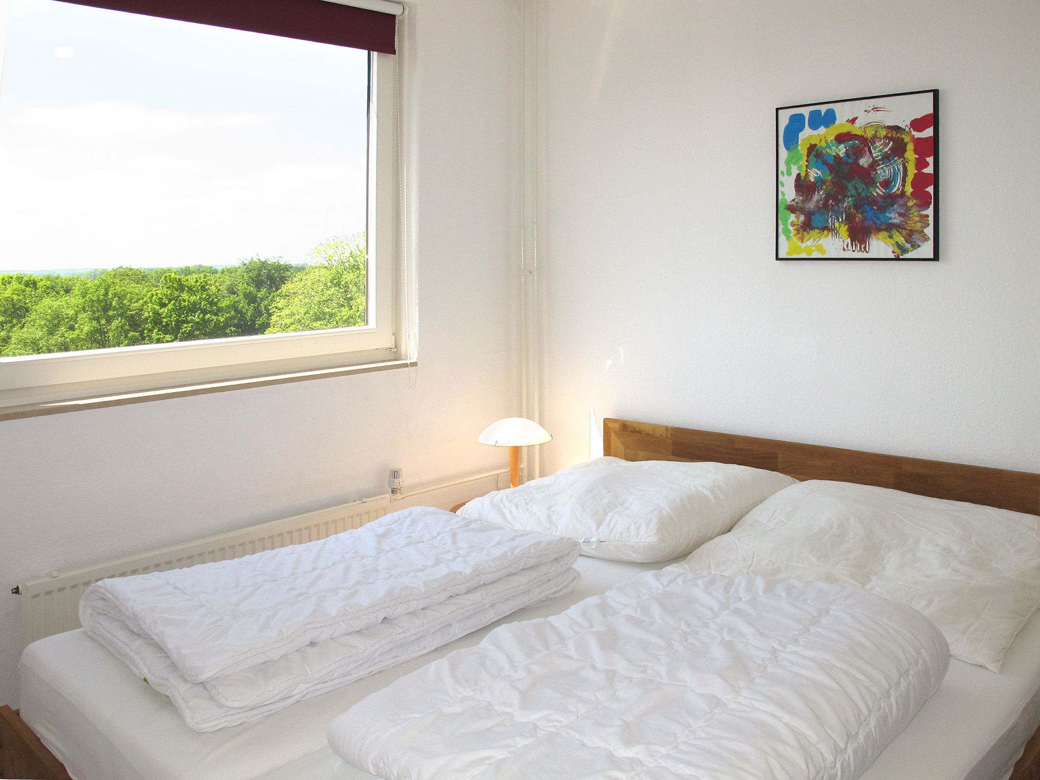 A neatly made bed with white bedding, a window with a view of greenery, and a colorful abstract painting on the wall.