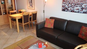 Una sala de estar con un sofá de cuero negro, una mesa de comedor de madera con sillas, una pequeña mesa de centro con una caja roja y papeles, una lámpara de pared y un cuadro de flores.